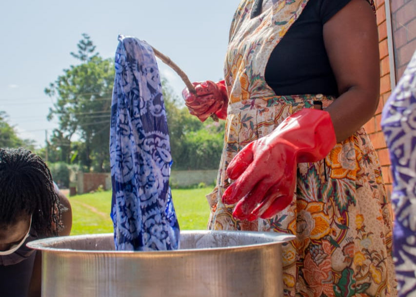 Woman wearing a face mask and gloves, standing next to a large metal pot outdoors makig batik in Uganda